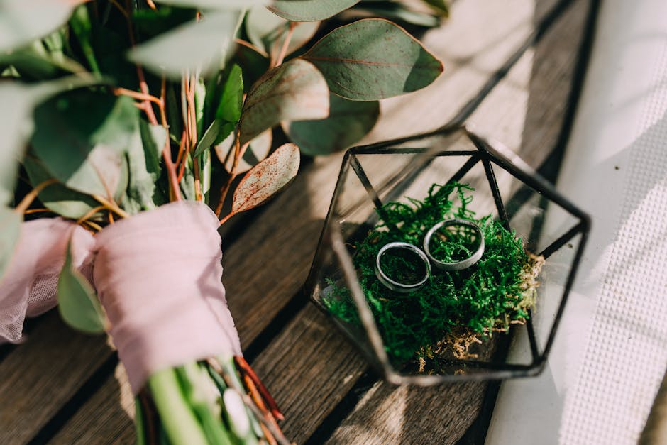 A close-up of a bouquet featuring eucalyptus leaves with silvery-green, rounded and elongated foliage, wrapped in soft pink paper tied with a pink ribbon, placed on a wooden surface. Next to it, a geo