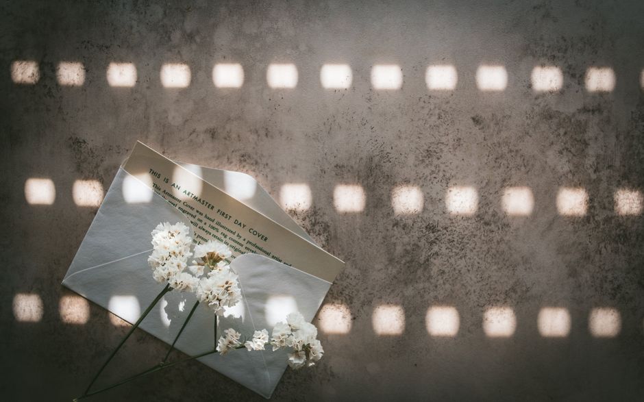 A white envelope labeled 'This is an Artmaster First Day Cover' is partially opened on a textured, grey concrete surface. A sprig of white flowers with delicate petals and green stems rests on top of