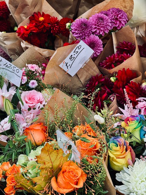 A vibrant assortment of freshly arranged flowers displayed at a market stall, featuring bright pink chrysanthemums, deep red dahlias, soft pink roses, orange roses, rainbow-colored roses, and white bl