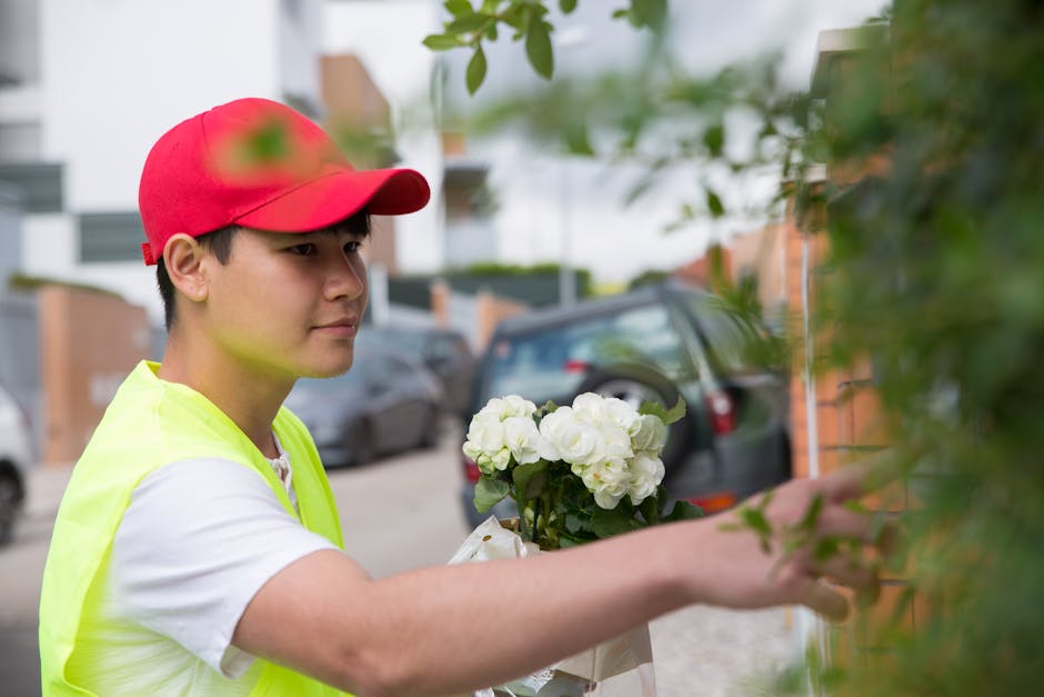 A young delivery person with fair skin, wearing a red cap and a neon yellow high-visibility vest over a white T-shirt, is outdoors extending her arm to place a bouquet of white flowers, possibly hydra