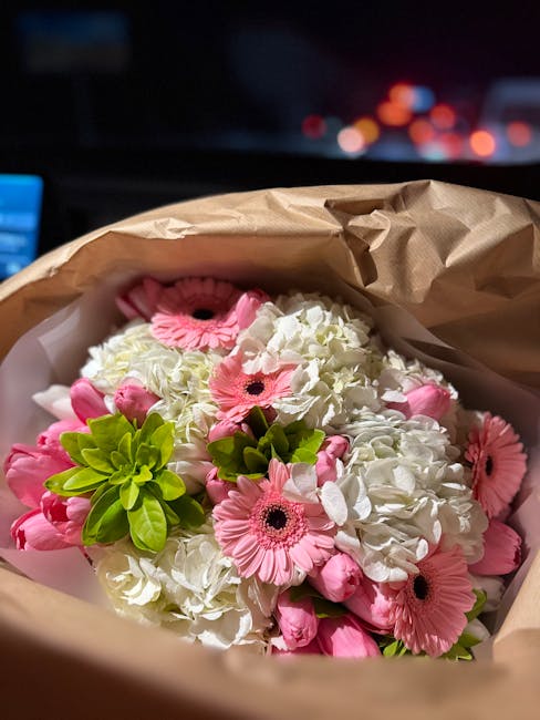 A bouquet of fresh flowers featuring white hydrangeas, pink gerbera daisies, and small green foliage, wrapped in light brown kraft paper with some white paper lining visible inside. The arrangement re