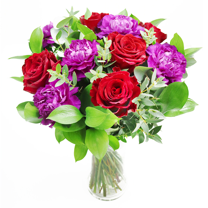 A beautifully rounded bouquet sits in a clear glass vase, looking both timeless and contemporary-very much at home in the creative spirit of Lower Clapton. The eye is first drawn to the deep red roses, each bloom velvety and full, its petals layering in tight yet generous spirals that radiate warmth and passion. Intertwined with them, purple peonies unfurl in sumptuous ruffles, their petals slightly rippled at the edges, creating a soft, billowing texture that contrasts with the more structured roses. The colour palette is rich and confident: crimson and purple blending like brushstrokes in a painting, each shade intensifying the other. A halo of fresh green foliage surrounds the flowers, the leaves broad, lush, and slightly overlapping, suggesting movement as though the bouquet has just been gathered from a thriving garden near Clapton Pond. Tendrils of eucalyptus meander through the arrangement, their rounded, silvery-green leaves introducing a cool note that keeps the design feeling light and contemporary. In the transparent vase, the stems are clearly visible, cut at considered angles and arranged in a gentle spiral, the different greens creating a subtle pattern through the water. The lighting appears natural, perhaps from a nearby window, with highlights on the rose petals and gentle shadows among the leaves, giving the scene depth and realism. In your mind, you might catch the layered scent: the enveloping sweetness of rose, the fresh lift of peonies just opened, and the clean, herbal hint of eucalyptus. This bouquet feels perfect for anniversaries, declarations of admiration, or adding a dramatic, romantic accent to a dinner table in a Lower Clapton flat or café.