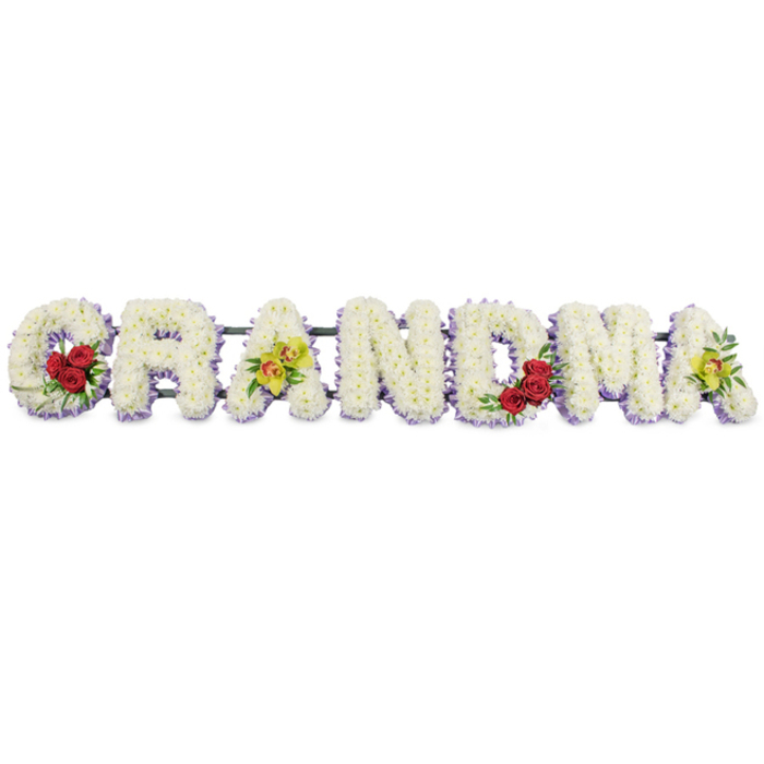 Spelling out the word GRANDMA in full capital letters, this floral tribute stretches gracefully across a bright, plain white background, allowing every detail of the craftsmanship to stand out. Each letter is meticulously filled with tight clusters of white chrysanthemums, their many-petalled heads pressed closely together to form a smooth, pillowed surface with a gentle sheen. Running along the lower edge of each letter is a delicate fringe of pale lavender blooms, adding a soft purple shadow that outlines and defines the shapes, like a quiet echo beneath the white. At the start of the word, the 'G' is adorned with three velvety deep red roses positioned at its lower left curve, framed by dark green leaves that provide rich contrast and signify deep respect and enduring affection. The first 'A' features two chartreuse orchids resting along its crossbar, their yellow-green petals and reddish-brown centres bringing an exotic, characterful note to the design. The central letters 'R', 'N', and 'D' are composed solely of the white and lavender blooms, their simplicity evoking calm reflection. On the 'M', a further trio of red roses nestles into the lower right section, visually balancing the roses on the 'G', while the final 'A' repeats the pair of fresh yellow-green orchids, giving a sense of completion and harmony. A slender dark metal frame discreetly supports the letters from above, making the flowers appear to hover against the white backdrop. This kind of GRANDMA tribute is often chosen by families in Lower Clapton and around Hackney to sit beside the coffin or at the front of a chapel, offering a tender, visually striking expression of love, memory, and thanks for a grandmother's life.